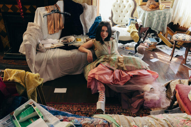 Singer Songwriter Mitski wears ornate vintage dress while sitting in densely decorated room