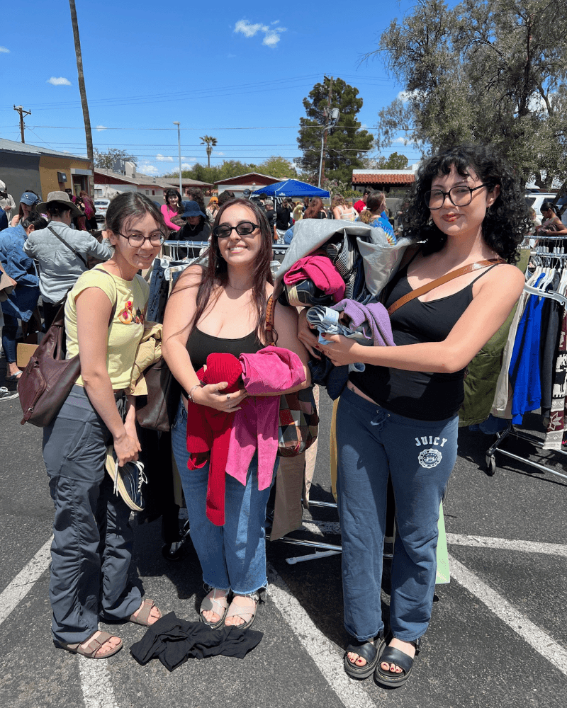 three women standing in a parking lot hold arms full of clothing